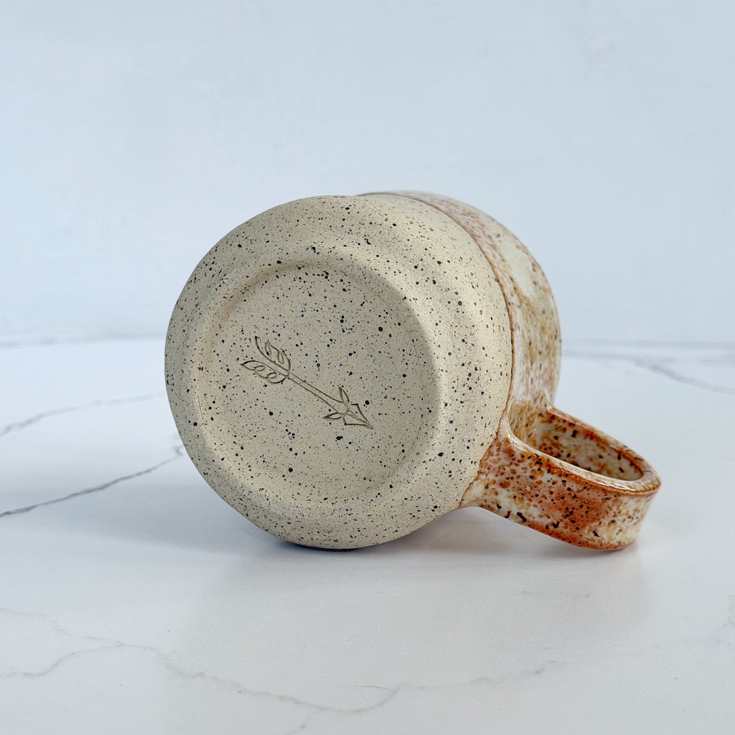 A handmade ceramic mug by Kathleen Kalish Ceramics with a speckled beige and brown glaze, shown from a side angle to show the potter's mark on a white marble surface against a light gray background.