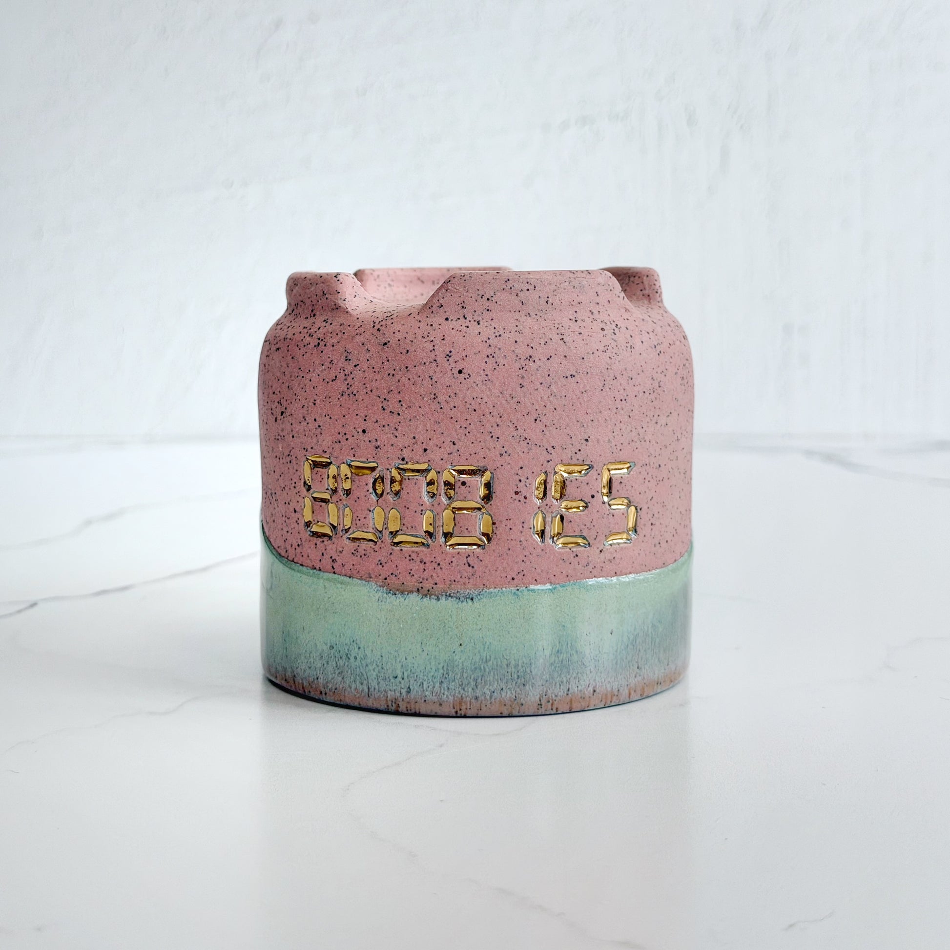 A handmade pink speckled clay ceramic cup by Kathleen Kalish Ceramics with green glaze. Stamped with BOOBIES and decorated with gold luster. For the Boobless Project helping breast cancer.