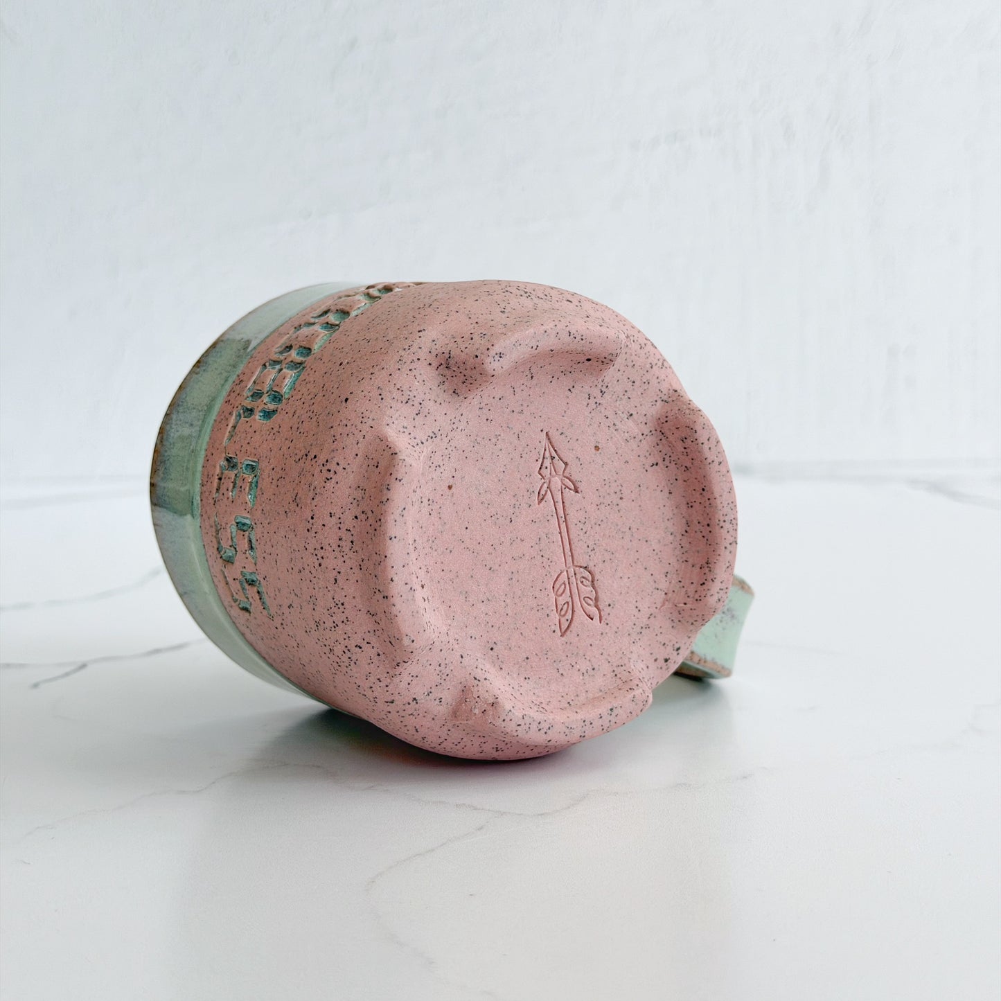 A handmade ceramic mug by Kathleen Kalish Ceramics with a speckled pink glaze and a mint green rim, shown lying on its side to display the bottom design featuring an embossed arrow and text.