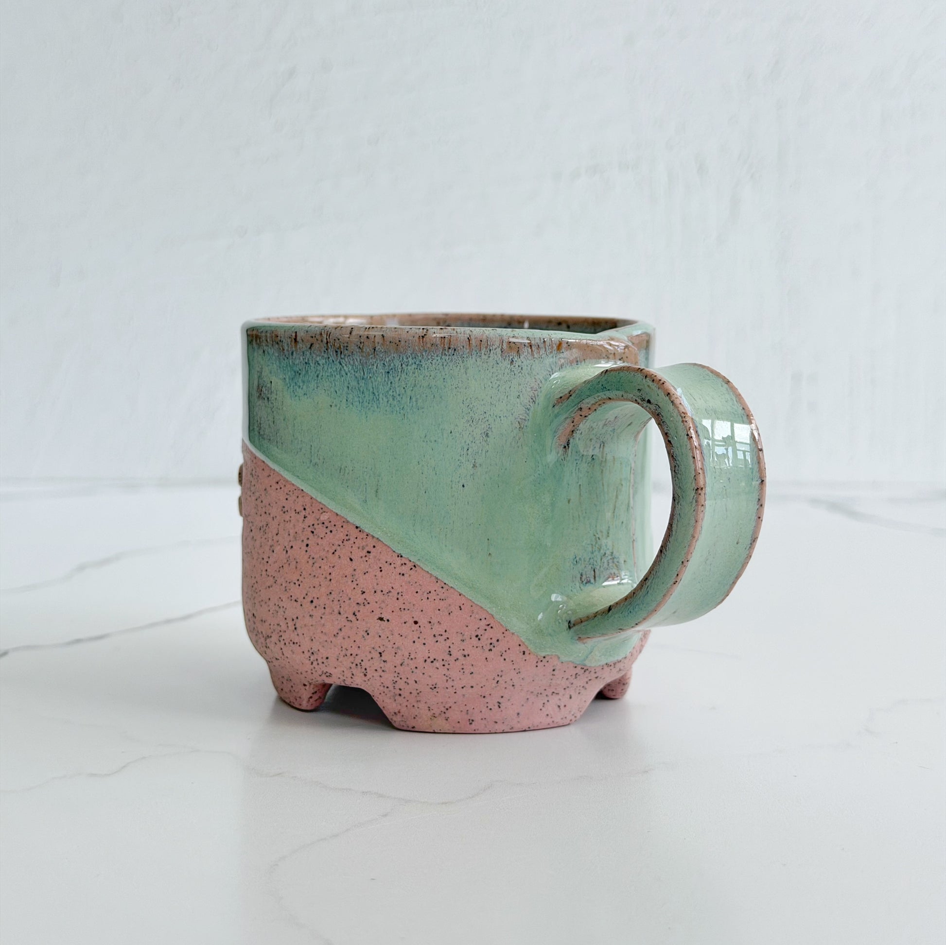 A handmade ceramic mug by Kathleen Kalish Ceramics with a diagonal split design, featuring a mint green glazed upper half and a speckled pink lower half, set against a light gray background.