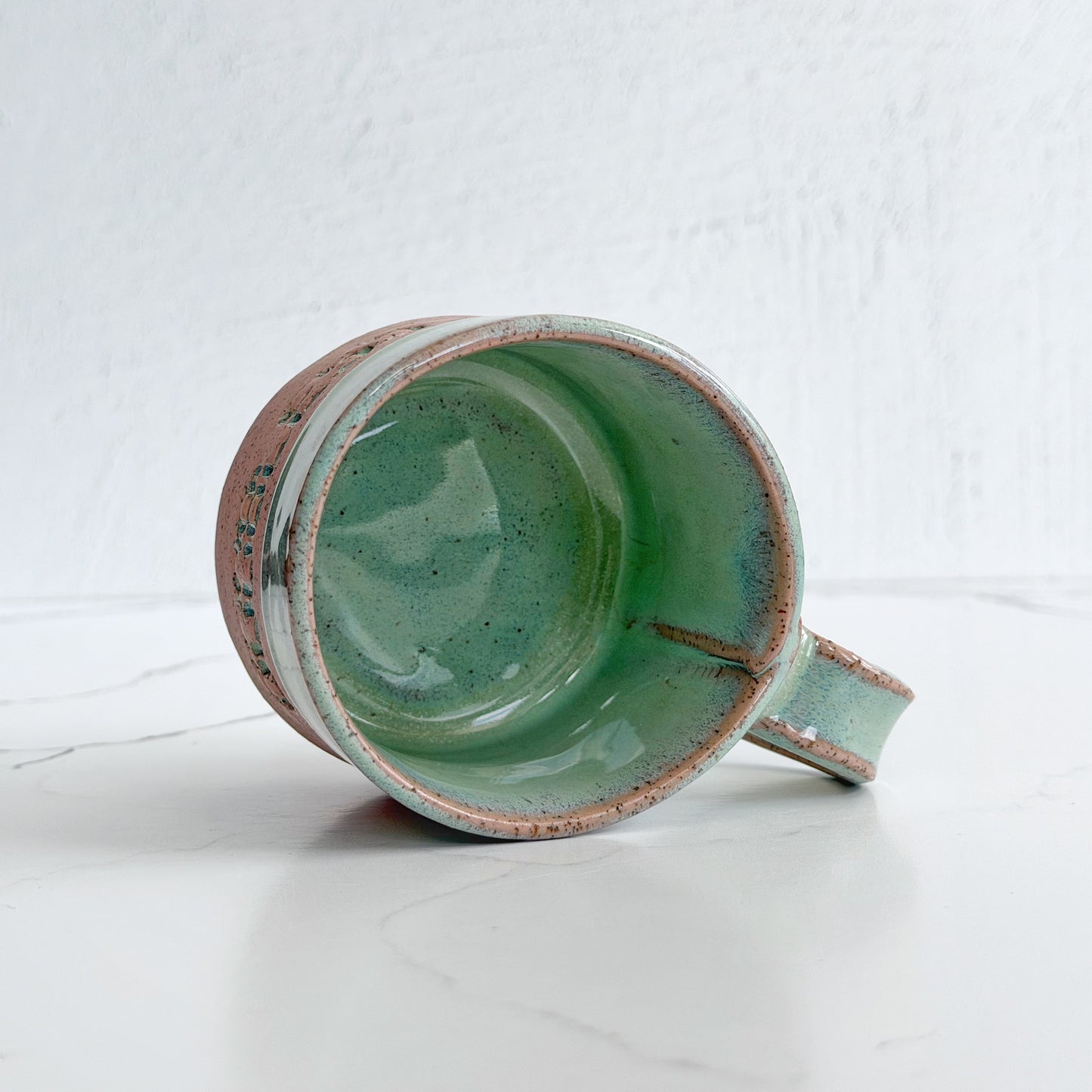 A handmade ceramic mug by Kathleen Kalish Ceramics with a glossy mint green glaze interior on pink speckled clay, shown lying on its side on a white marble surface against a white textured wall.