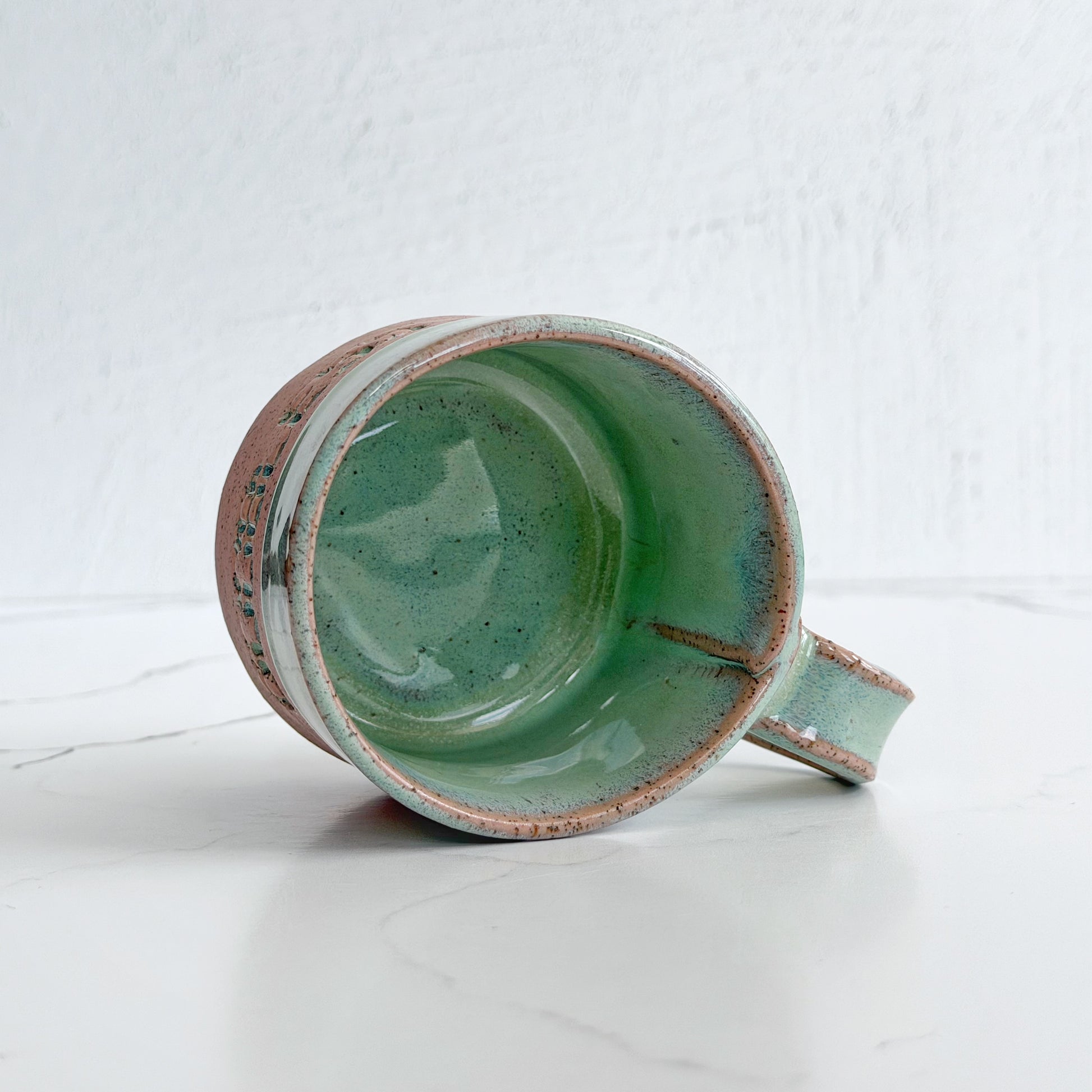 A handmade ceramic mug by Kathleen Kalish Ceramics with a glossy mint green glaze interior on pink speckled clay, shown lying on its side on a white marble surface against a white textured wall.