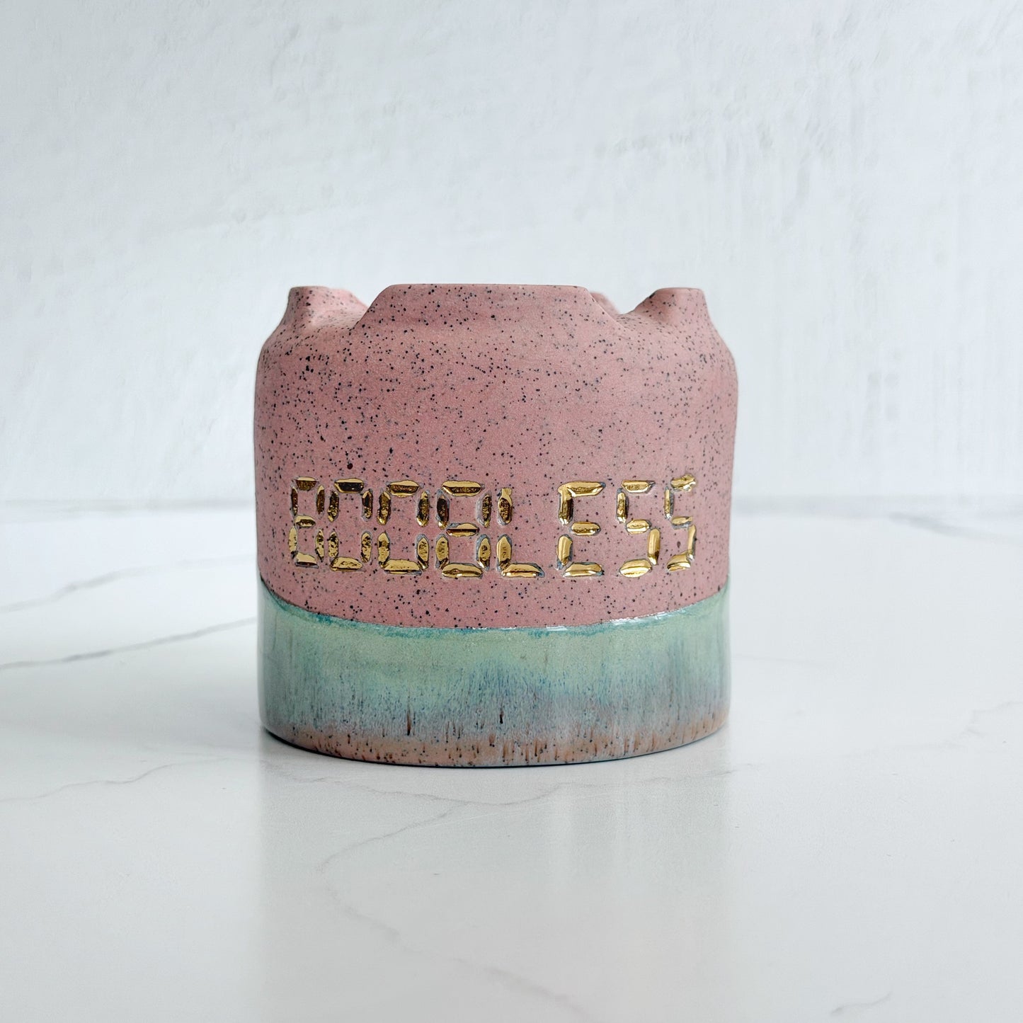 A handmade pink speckled clay ceramic cup by Kathleen Kalish Ceramics with green glaze. Stamped with BOOBLESS and decorated with gold luster. For the Boobless Project helping breast cancer.