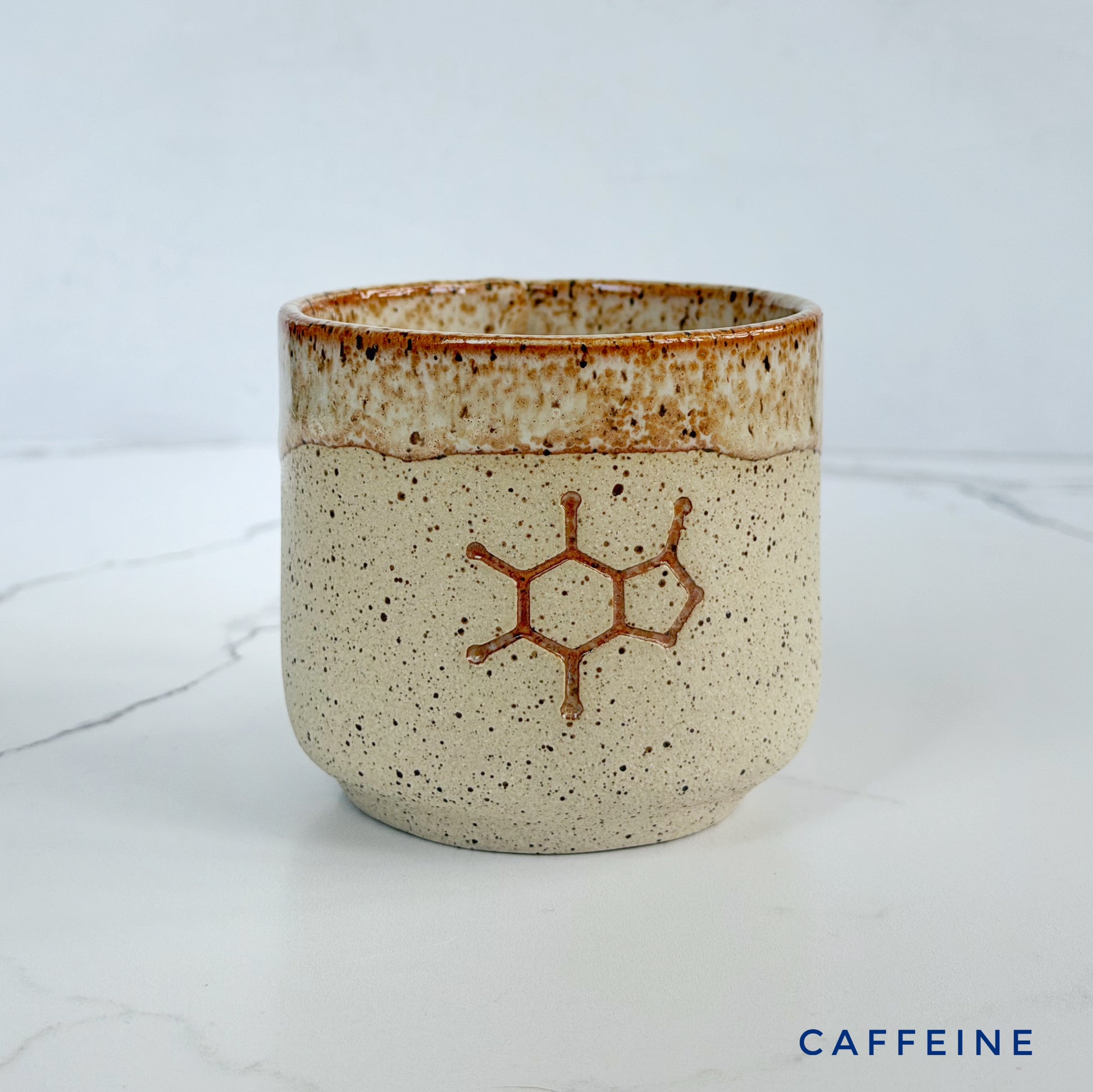 A handmade ceramic coffee cup by Kathleen Kalish Ceramics with a speckled beige and brown glaze, featuring a brown design of the caffeine molecule on the front. The cup is set on a white marble surface against a light gray background.
