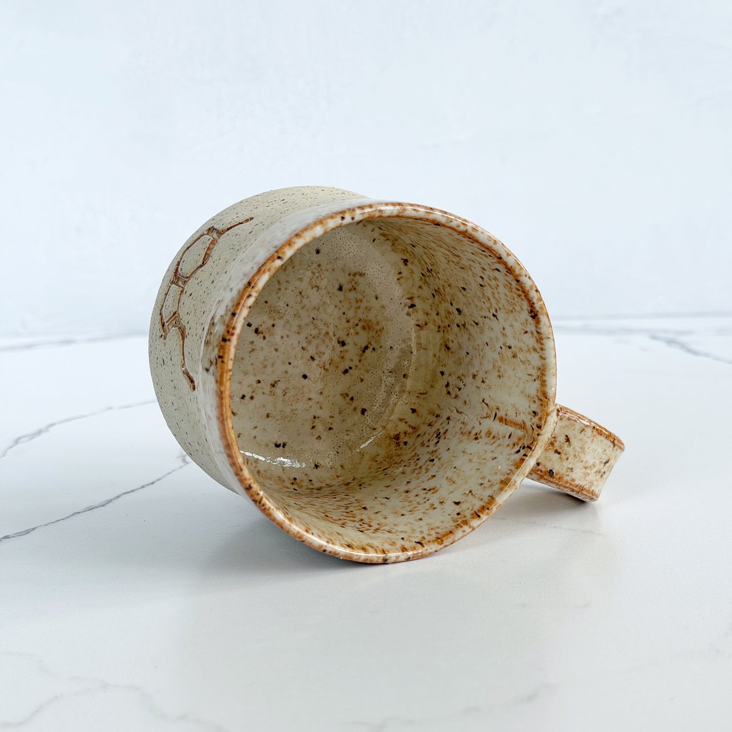 A handmade ceramic mug by Kathleen Kalish Ceramics with a textured, speckled beige and brown glaze, shown lying on its side on a white marble surface against a white background.