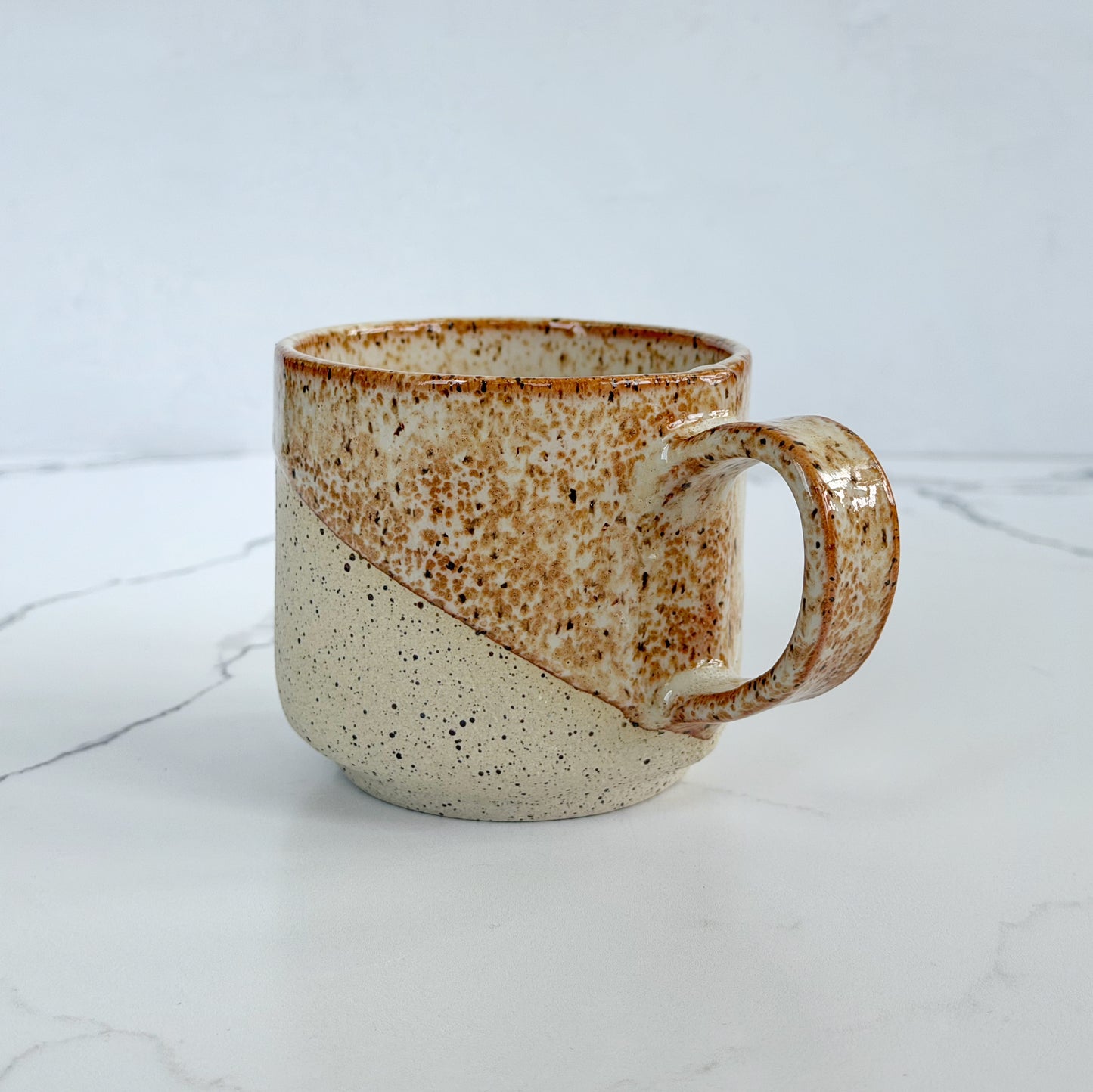 A handmade ceramic mug by Kathleen Kalish Ceramics with a diagonal split design, featuring a speckled brown and beige top half and a cream-colored bottom half with dark speckles, set on a white marble surface.