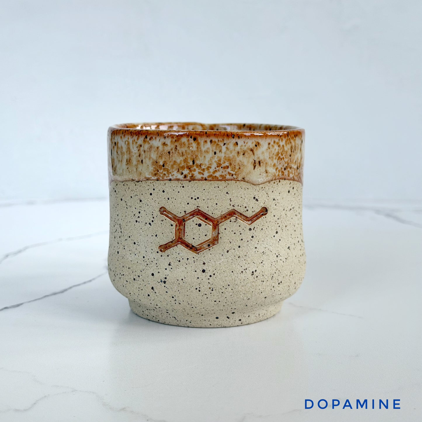 A handmade ceramic cup by Kathleen Kalish Ceramics with a speckled beige body, a brown glazed rim, and a brown dopamine molecule design on the front, set on a white marble surface against a light gray background.