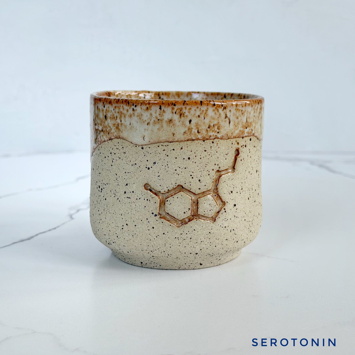 A handmade ceramic cup by Kathleen Kalish Ceramics with a textured speckled beige body, a brown glazed rim, and an embossed serotonin molecule design on the front, set against a light gray marble background.